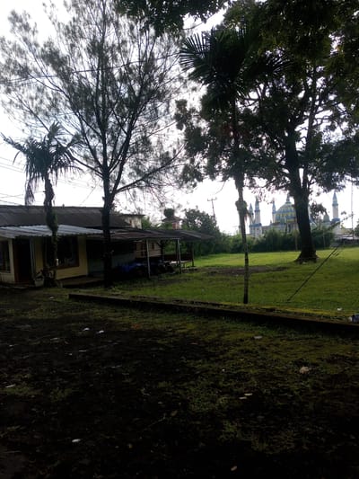 The image shows a mosque in the distance, with a foreground containing a building and trees. The mosque has a blue and yellow dome and minarets. A two-section, one-story building with a corrugated metal roof is on the left side of the scene. Trees partially obscure the mosque. The foreground contains what appears to be dirt, grass, and some trash. The image may have been taken in Mya Sit Kaing Ywar Thit, Myanmar.