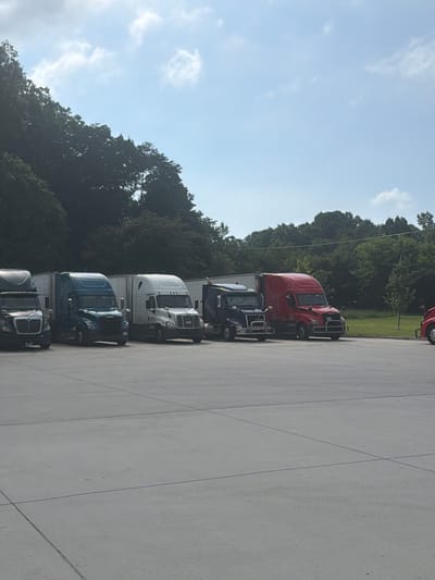 The image shows a row of parked semi-trucks on a paved lot. The trucks are of various colors, including black, teal, white, and red. Some of the trucks have trailers attached. In the background, there is a dense line of trees and a blue sky with some clouds. The scene appears to be a truck stop or a logistics yard. The location is not identifiable within the city of Chattanooga, United States of America, and no people are visible in the image.