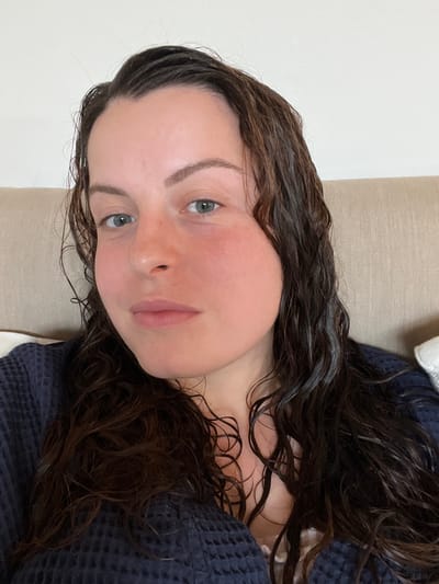 A close-up, head-and-shoulders portrait depicts a female subject in an indoor environment. The subject has fair skin, light-colored eyes (appears blue-grey), and dark brown, wavy hair that appears wet or damp, extending over her shoulders and chest. Her cheeks and nose exhibit a flushed appearance. She is looking directly at the camera with a neutral expression. She is wearing a dark blue, possibly navy, waffle-knit robe or similar garment, with a lighter-colored undergarment visible at the neckline. The background consists of a light-colored, textured fabric, likely a headboard or couch back, with a partial view of a white pillow to the right. The scene is located in Bad Sobernheim, Germany.