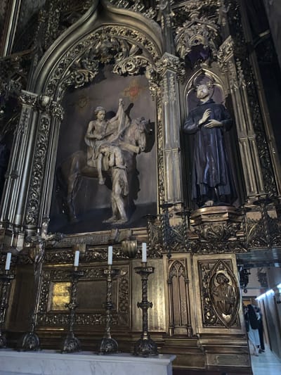 The image depicts the interior of a richly decorated church in Monistrol de Montserrat, Spain, likely the Monastery of Montserrat. In the center, a large, ornate gilded altarpiece dominates the scene. To the left, a light-colored sculptural group features a man on horseback, with another figure in distress leaning towards the horse. To the right, a dark-robed statue of a saint stands on a pedestal, hands clasped. The altarpiece is adorned with intricate carvings, architectural elements, and decorative panels. Several tall, slender candlesticks with white candles are arranged in the foreground, resting on a marble surface. In the background, to the right, two individuals are visible, walking away from the camera, suggesting the space is open to visitors. The overall impression is one of historical and religious grandeur, with meticulous craftsmanship evident in every detail.