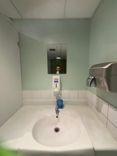 The image shows a bathroom sink in a public restroom. The sink is made of white porcelain and has a chrome faucet. There is a hand sanitizer dispenser next to the faucet and a hand dryer on the right side of the sink. The wall tiles are white and the walls are painted a light green. There is a mirror above the sink that reflects a person using a cell phone. The coordinates 53.794, -1.752 correspond to Bradford, United Kingdom, and based on the architecture and style of the room, this is likely a public restroom within an office building or other commercial space in the city of Bradford.