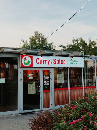 This media file features the exterior of an Indian restaurant named "Curry & Spice" in Nowy Dwór Mazowiecki, Poland. The restaurant has large glass doors and windows, with the name "Curry & Spice" prominently displayed in red lettering above the entrance, accompanied by a logo featuring a chili pepper and a spiral. To the right of the restaurant name, it is identified as an "INDYJSKA RESTAURACJA" (Indian Restaurant).
Visible on the glass doors are various signs, including operating hours ("GODZINY OTWARCIA"), stating the restaurant is open "Cały Tydzień" (All Week) from "11:00 do 22:00" (11:00 AM to 10:00 PM), and a phone number "+48 888 070 085". A "Lunch Menu" is also displayed. Inside the restaurant, through the windows, people can be seen seated at tables, suggesting the restaurant is open for business.
The setting appears to be outdoors, with greenery and flowering bushes in the foreground. The lighting suggests it is either early morning or late afternoon, with a soft, natural light. The sky is visible and appears clear. The overall scene conveys a casual and inviting atmosphere for dining.