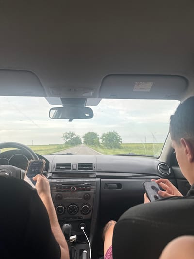 The image provides an interior view of a car, oriented as if looking forward from a rear seat, set in Topraisar, Romania.
Two individuals are visible. The person in the driver's seat has their arms on the steering wheel, with their right hand interacting with a smartphone that appears to be mounted or placed near the wheel. Their head and upper body are partially visible. In the front passenger seat, another person's upper body is seen, actively holding and looking at their own smartphone.
The car's interior features a dashboard with air vents, a central console containing an audio system with radio/CD player controls, and climate control dials (AC, fan speed). A gear shifter, a white charging cable, and a rearview mirror are also visible. The driver's sun visor is down, showing a safety warning label.
Through the windshield, a rural landscape is visible, characterized by green fields and several distant trees. The road ahead appears to be a paved path through this natural environment. The sky is bright but overcast, suggesting a cloudy daytime.
Both the driver and the passenger are engaged with their mobile phones. The driver's phone screen shows what looks like a music player interface or navigation, while the passenger is focused on their device.
Visible text includes:
* On the driver's sun visor: A "WARNING" label with a child safety symbol.
* On the central console: "FM/AM", "DISC", "CD", "AUX", "MODE", "AUTO", "AC", and the "HAZARD" button.
* On both phones: Various app icons are discernible on their screens.