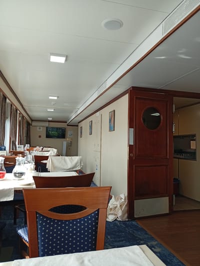 The image shows a dining area on a ship, possibly a river cruise ship, in Skipino (Timiryazevskii S/S), Russia. The room is long and narrow, with tables set for dining. The tables are covered with white cloths and have chairs with dark blue patterned upholstery. There are pitchers of water and other drinks on the tables, along with glassware. Windows with brown curtains are visible on the left side of the image.

On the right side, there is a wall with light beige panels, some with framed artwork. A wooden door with a circular window leads to what appears to be the ship's kitchen or service area, with stainless steel equipment visible inside. A stack of white cloths is visible on the floor near the door. The floor has a blue carpet in the dining area and wooden flooring in the kitchen/service area.

The ceiling is white with recessed lighting and ventilation grilles. The overall impression is of a well-maintained dining area on a passenger ship.