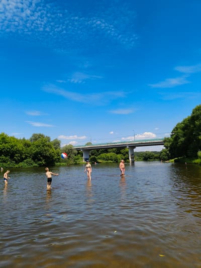 In the city of Uspenskoye, Russia, on a clear summer day, several people are enjoying the water of a wide river. A multi-colored inflatable ball is in mid-air, suggesting a game is in progress. Two young boys are wading in the shallow water, one in blue swim trunks, the other in black shorts. A young woman, wearing a bikini, stands facing the boys. Further in the distance, a man in patterned swim trunks is walking through the water. The river is surrounded by lush green trees on both banks, and a modern concrete bridge with a light blue railing spans the water in the background. The sky is a vibrant blue with scattered white clouds.