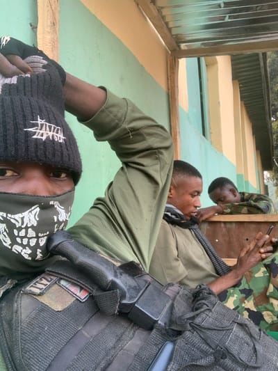 Three people are shown in the image. One person is wearing a black beanie and a bandana over the lower half of their face. To their right, a person in a tan shirt and scarf looks up. Next to them, another person in camouflage holds a cell phone to their ear. The people are in front of a tan and green painted wall. The person closest to the camera wears a tactical vest with a holster and magazine pouch.
