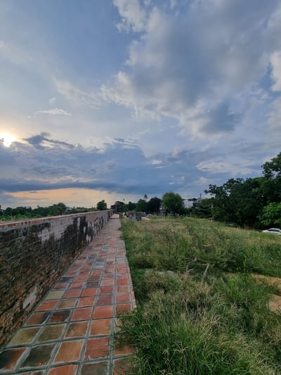 The image shows a tiled pathway along an ancient brick wall. The wall is on the left side of the frame, showing signs of age and wear. The pathway is made of square terracotta tiles laid in a diamond pattern and extends into the distance. To the right of the pathway, there is a field of overgrown grass and some trees. In the background, there are buildings and more trees, suggesting an urban or semi-urban environment. The sky above is cloudy with patches of blue sky and the sun is partially visible through the clouds on the left side of the frame, indicating either sunrise or sunset. The location is identified as Phú Xuân Ward, Vietnam. No people are clearly visible, though a few distant figures may be present in the background. A white car is parked on the right side of the frame, near the trees.