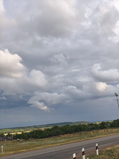 The image depicts a landscape scene with a dramatic sky filled with heavy, gray clouds. A paved road runs through the foreground, bordered by dry grass and low vegetation. In the midground, there is a line of trees and rolling hills with scattered houses and fields. A street lamp is visible on the right side of the image, and power lines stretch across the horizon. The overall impression is one of a potentially stormy or overcast day in a rural or semi-rural setting. The location is specified as Rodionov, Russia.