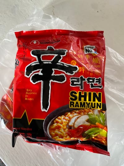 A red package of Nongshim Shin Ramyun instant noodles. The packaging includes the brand name, "Shin Ramyun" in prominent gold lettering, along with Korean text. A vibrant image shows a bowl of noodles with toppings. Text proclaims "Korea No. 1" and "Spicy Happiness in Noodles." An egg logo is displayed alongside "Lovely Life good food." The expiration date on the package is "OCT-2025." Net contents are listed as "120g."
