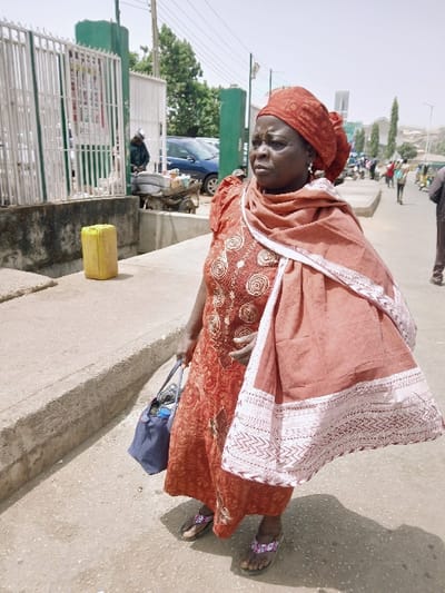 The scene depicts an outdoor urban environment in Kadoma, Zimbabwe. A dark-skinned adult female stands centrally on a light-colored paved surface, looking slightly to her left. She wears a reddish-orange patterned long dress, a matching headwrap, and a reddish-orange shawl with a white patterned border draped over her left shoulder. Her left hand holds a dark blue tote bag, and she wears purple and white patterned flip-flops. To her left, a concrete curb runs horizontally, separating the paved area from a lower ground section where a yellow plastic jerrycan is present. Behind this, a white metal fence with green posts encloses an area where a person in dark clothing is visible next to a two-wheeled cart laden with goods. A blue car and other vehicles are parked further back. The background shows a continuation of the paved area with multiple people walking or standing, and distant buildings on elevated terrain under a bright, clear sky. Shadows are cast on the ground, indicating direct sunlight.