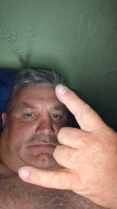 A close-up, selfie-style photograph shows a man with graying hair and blue eyes. He is looking directly at the camera. His right hand is raised, with the index finger and pinky extended in a gesture commonly associated with rock and roll. The background is a textured, green wall. The image appears to have been taken indoors. There is no information within the image to indicate the location is Voinova, Moldova.