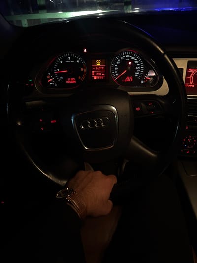 The image shows the interior of an Audi car, focusing on the steering wheel and dashboard. The car's instrument panel displays a temperature of 14.0°C and a mileage reading of 230 km. The speedometer indicates a speed of 0 km/h, and the tachometer reads approximately 2150 RPM. The date on the display is 17.05.2026. A person's hand wearing a watch is visible on the right side of the steering wheel. The background is dark, suggesting a night time setting. There is no indication of the location being Valchedram, Bulgaria.