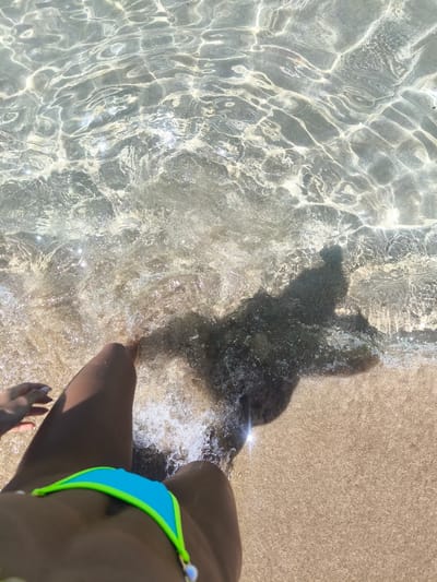 A high-angle shot captures the edge of shallow, clear ocean water lapping onto a sandy beach. The sun reflects off the water's surface, creating shimmering patterns. A person's legs, wearing a turquoise and lime green bikini, are visible in the foreground, with their left hand reaching towards the water. A dark shadow of a person is cast on the seabed. The image appears to be taken at a beach in Paphos Municipality, Cyprus.