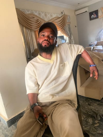 An adult male is seated indoors, likely within a residence in Eika-Adagu New Layout, Nigeria. He is positioned facing the camera, wearing a black baseball cap, a cream-colored V-neck short-sleeved top with a lighter yellow trim and a left chest pocket featuring a small metallic emblem, and light brown trousers. He has a full beard and is looking directly forward. A black wristwatch is present on his left wrist, and a blue silicone wristband with "Bungie" inscribed in white text is on his right wrist. His left hand holds a black feature phone. He is seated on a chair with a dark-colored back, positioned in front of a light cream-colored wall. The background features light brown/gold patterned curtains, an air conditioning unit mounted on the wall to the right, and a framed portrait below the AC unit. A beige upholstered sofa is partially visible behind him to the right. The floor is dark patterned marble or tile.