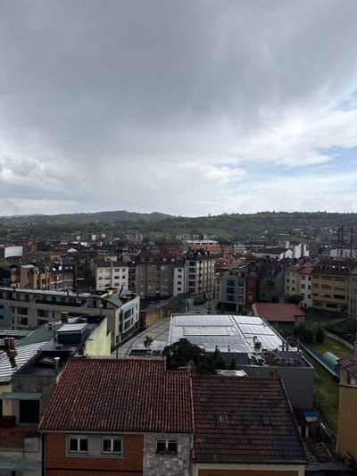 The image is a high-angle, slightly tilted, daytime shot of the city of Oviedo, also known as Uviéu, in Spain. The sky is overcast with mostly grey clouds. Below, a dense urban landscape is visible, featuring a multitude of buildings of varying heights, designs, and colors. Many structures have tiled roofs, predominantly in shades of red and brown. Some modern buildings with flat roofs and metallic or glass surfaces can also be observed.

In the background, a green hillside rises, providing a natural boundary to the urban sprawl. The buildings are densely packed together, showing a typical European cityscape. No specific people are visible. The environment appears to be wet, possibly due to recent rain.