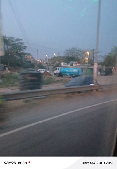 A blurry, low-light image captures a roadside scene from a moving vehicle. A road with a white dividing line occupies the foreground. Beyond a guardrail, various objects appear: a large, dark storage tank, a mound of earth, a blue water truck, and parked cars. Trees and streetlights line the distant background under a dim sky. The camera settings, noted at the bottom, suggest a mobile phone shot with minimal stabilization.