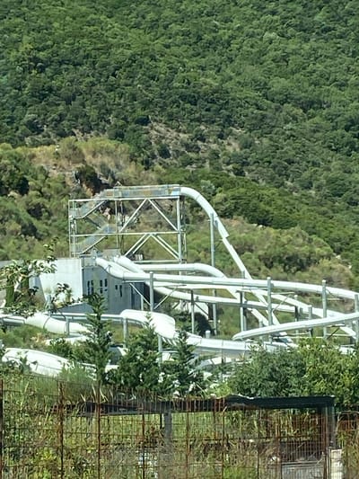 The image depicts an abandoned water park situated in the city of Seoce, Montenegro. The water park, characterized by its white plastic slides and metal support structures, is partially overgrown with vegetation. In the background, a dense forest covers a hillside. The scene suggests a state of disrepair, with the slides and structures appearing weathered and unused. There are no people visible in the image.