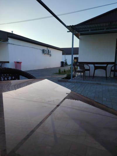 The image depicts an outdoor seating area adjacent to a white building with a dark-pitched roof. Two air conditioning units are mounted on the exterior wall of the building. In the foreground, a reflective, light-colored surface, likely a table, occupies the majority of the frame. A dark wicker chair is partially visible to the left. The ground is paved with interlocking gray bricks. In the background, another white building with a similar dark roof is visible, with a pink trash can and dark fencing. The sky is clear and light blue, suggesting it is daytime, possibly late afternoon or early morning due to the lighting. The setting appears to be a commercial or residential complex.