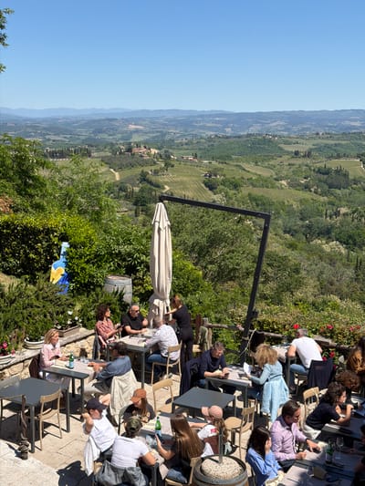 The image presents a vibrant outdoor dining or social gathering scene on a patio overlooking the panoramic Tuscan countryside, identified as being near San Gimignano, Italy. Numerous adults are seated at rectangular tables or standing, engaged in conversation, dining, and drinking. Many are dressed in casual to smart-casual attire. Tables are equipped with glasses and bottles, some clearly identifiable as green San Pellegrino water bottles.

The patio itself is paved and features a large, light-colored patio umbrella offering shade, as well as a prominent black metal structure, possibly part of an awning or architectural element. Lush green bushes, flowering plants with red and white blossoms, and a distinctive large blue and yellow ceramic vase or sculpture adorn the edges of the dining area. A wooden barrel filled with small stones or gravel is also visible on the patio.

Beyond the immediate patio, a vast and picturesque landscape unfolds under a clear, bright blue sky. Rolling green hills covered in vineyards, dense forests, and scattered traditional Italian buildings forming distant villages characterize the scenic view. The bright sunlight and strong shadows suggest the time of day is midday or early afternoon, indicating pleasant, clear weather. The overall atmosphere appears relaxed and social, with people enjoying the company, refreshments, and the stunning natural beauty of the Italian countryside. No discernible text is visible within the image.