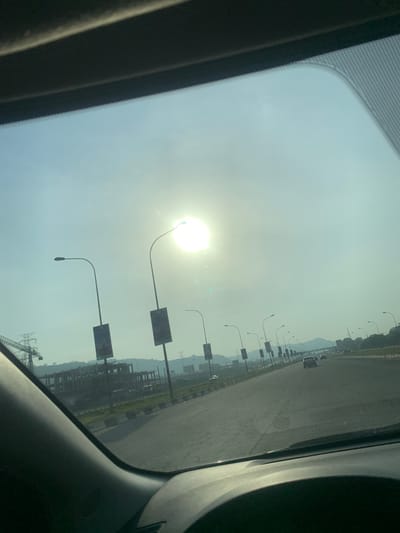 View from inside a car through the sunroof. The sun is high in the sky, blurring details. A paved road stretches into the distance lined with street lamps and signage. Construction is visible on the right side of the road. A mountain range is visible further in the distance.