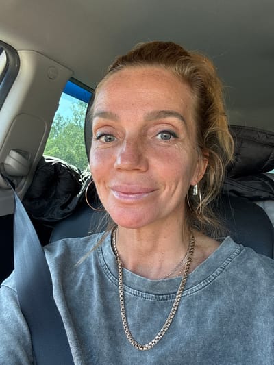 The image depicts a woman seated inside a vehicle. The stated location is Beloostrov, Russia, although this detail is not discernible from the visual content. She is positioned in what appears to be the driver's seat, wearing a grey seatbelt diagonally across her left shoulder and chest. The woman has light brown hair, pulled back, and a fair complexion with visible freckles across her nose and cheeks, along with some sun-induced redness. Her eyes are blue-grey, with visible dark eyeliner on the lower lid and mascara. She is looking directly at the camera with a subtle, closed-mouth smile.

She wears two gold-toned chain necklaces, one thicker with a rectangular link pattern and one thinner. Her attire consists of a grey, short-sleeved t-shirt with a visibly distressed or stonewashed texture. She has two different earrings: a large, thin, gold-toned hoop on her left ear, and a smaller, textured, silver-toned hoop with an additional small charm on her right ear.

The vehicle's interior features light grey overhead lining and pillar trim, with an "AIRBAG" indicator visible on the A-pillar. A black seat and a black puffy jacket or bag are visible on the passenger side. Through the vehicle's windows, green foliage from trees and a clear blue sky are discernible, indicating an outdoor, daytime setting.