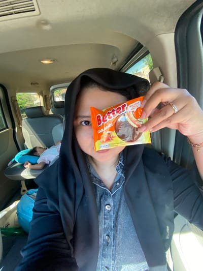 A selfie taken from inside a vehicle shows a woman in a black hijab and denim shirt holding a package of "Better" caramel biscuits in front of her face, partially obscuring it. To her left, a young child sleeps on a blue pillow in the car's back seat. The car's interior is visible, including the ceiling, air vents, and seats. The background shows greenery and buildings, suggesting an outdoor location. The scene is likely taking place in Randubango, Indonesia.