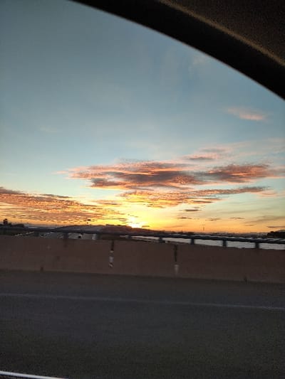 A vibrant sunset unfolds through a car window. Fiery orange and red clouds streak across a light blue sky. A bridge or barrier lines the horizon, beyond which lies a strip of land. The foreground is dominated by the interior of the car, with a dark frame partially obscuring the view. A highway with marked lines stretches to the right.