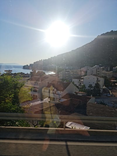 This image depicts a sun-drenched coastal town, likely Drvenik, Croatia, with a prominent view of the Adriatic Sea. The sun is intensely bright, creating lens flare and scattering light across the scene. The town features a collection of white and light-colored buildings, many with tiled roofs, nestled on a hillside that slopes down towards the water. The architecture suggests a Mediterranean style, with multi-story apartment buildings and smaller residential houses visible.
The foreground is dominated by a concrete barrier and railing, indicating the viewpoint is likely from a road or elevated walkway. Green foliage from trees and bushes partially obscures the view of the buildings in the lower left portion of the frame.
The sea stretches out to the horizon, appearing calm and reflecting the bright sunlight. In the distance, faint landmasses can be seen. The hillside behind the town is covered in trees, giving it a rugged, natural appearance.
Several cars are parked on a road winding through the town, and there are glimpses of streets and pathways between the buildings. The overall impression is of a serene, sunny day in a European coastal resort. There are no people clearly visible in the frame. The image appears to have been taken during the daytime, likely late morning or early afternoon, given the angle of the sun and the quality of light.