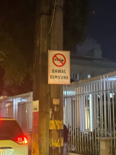 The image captures a street scene at night in Manila, Philippines. The primary focus is a vertical sign on a pole, depicting a red circle with a diagonal line over a horn icon, indicating "No Honking." Below the icon, text in Filipino reads "BAWAL BUMUSINA," which translates to "Honking is Forbidden." The sign is bordered in red and white.

In the background, a modern white structure with horizontal slats is visible, possibly part of an overpass or building. The lighting suggests it's nighttime, with artificial lights from the street and possibly from vehicles illuminating the scene. Hints of greenery are present at the bottom, suggesting trees or bushes. A small, blurry detail in the upper left corner shows two colored lights in the dark sky, which could be distant lights or reflections.

Further down the pole, other faded signs or stickers are partially visible. To the right of the pole, a portion of a vehicle's taillight is illuminated, showing red brake lights, indicating a vehicle is stopped or moving slowly. The overall atmosphere is urban and nocturnal. There are no people clearly visible in the frame. The weather appears to be clear, as there are no indications of rain or fog.