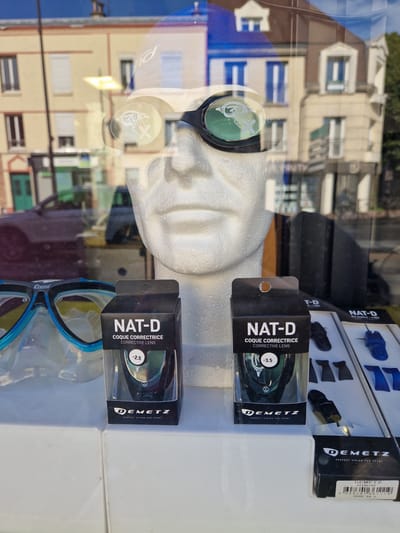 A shop window display in Antony, France, features a mannequin head wearing swimming goggles with corrective lenses. Two boxes of "NAT-D COQUE CORRECTRICE CORRECTIVE LENS" are positioned in front of the mannequin, one displaying "-2.5" and the other "-3.5" on their respective lenses. A blue and black swimming mask is also visible. The background reflects buildings and trees. The lower right corner of the image shows a barcode sticker for a Demetz product.