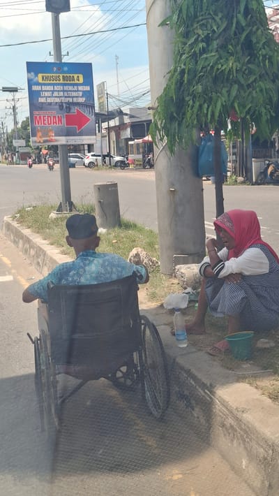 The image depicts a street scene in Pantegajah, Indonesia. In the foreground, a man in a wheelchair wearing a blue patterned shirt and a black cap is positioned on the left side of the frame. To his right, a woman in a red hijab and a striped dress is sitting on the curb, appearing to be speaking. A plastic water bottle and a green bucket are on the ground near her. Behind them, a large concrete utility pole stands with a blue plastic bag hanging from it. A road with traffic, including motorcycles and cars, is visible in the background. A prominent blue billboard with text in Indonesian and a red arrow pointing to the right is mounted on a pole on the left side of the road. The environment appears to be a sunny day with a clear sky.