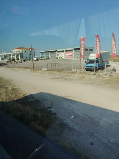 The image shows a light blue truck with a grey tarp and text in Greek on its side parked in front of a warehouse-like building. Three red flags with the white text "DAST" are planted in the ground near the building. To the left of the warehouse, a white building with a red-tiled roof and large glass windows is visible. There are also some metal fences and a dirt road in the foreground. The scene appears to be in a commercial or industrial area on a sunny day with a clear blue sky. The location is identified as the Municipal Unit of Makrichori, Greece.