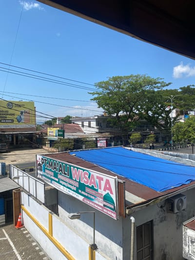 The image depicts a street scene in the city of Keraton, Indonesia, under a clear blue sky. In the foreground, a white wall with yellow accents runs horizontally, with an orange traffic cone placed on the paved ground to the left. A prominent sign for "PT. MA'ALI WISATA" is mounted on a railing above the wall, advertising "UMRAH PLUS," "UMRAH HAJI KHUSUS," and "UMRAH REGULER," along with a phone number. To the right of the wall, a building has a rusted tin roof covered with a blue tarp, and an air conditioning unit is visible on the exterior wall. Further back, various buildings with red tile roofs line the street, interspersed with lush green trees, including a large, mature tree dominating the midground. Power lines crisscross the sky. A yellow billboard with an image of a smartphone and the text "JORAGAN STORE" is visible on the left side of the frame. Several motorcycles and a black car are parked along the street. People are present but are not the primary focus. The overall impression is that of a busy, lived-in urban environment with commercial activity.