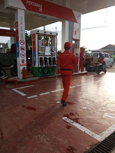 A gas station attendant in a red uniform and cap walks away from the camera on the wet, red-colored ground of a Pertamina gas station. The attendant is carrying a key in his right hand. In the background, a gas pump with multiple nozzles stands tall, with various signs and decals on its structure. A red fire extinguisher is visible on the side of the pump. To the left of the pump, a red truck is partially visible. In the far background, several cars and a motorcycle are parked, and two other individuals in red uniforms are standing near a silver car. The environment appears to be an outdoor, sunny location. The overall scene depicts a typical operational day at an Indonesian gas station.