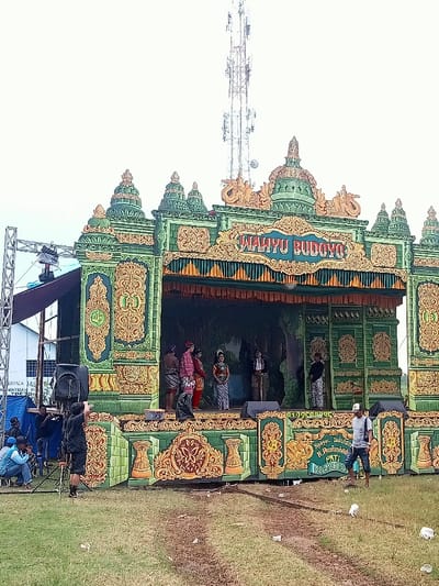 The image shows a stage decorated in an elaborate, ornate style with a green and gold color scheme. The stage is fronted by a sign that reads "Wahyu Budoyo," and it is located in Kecamatan Margorejo, Indonesia. The stage is set up in a grassy field, with some debris scattered on the ground. A tall, white radio tower with multiple antennas is visible in the background. Six people dressed in traditional clothing are standing on stage, facing forward towards the audience. Two men are standing off to the side of the stage; one is wearing a black shirt and holding a black camera, and the other is wearing a blue shirt and a black hat. A few other people are visible in the background, standing in the grass. There is a blue tarp and several chairs set up in the foreground. The scene is likely a performance or event, with the people on stage seemingly involved in a play or dance. The stage is built on a raised platform and appears to have a backdrop depicting a forest or jungle scene.