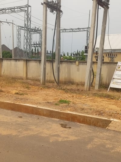The image depicts an outdoor scene at an electrical substation in Baccure Kofare, Nigeria. The foreground features a paved road with a concrete curb, and a patch of dry, uneven ground with sparse vegetation. Beyond this, a concrete wall forms a boundary. Behind the wall, a complex electrical substation is visible, with metal towers supporting high-voltage wires and insulators. Several large, grey concrete poles, likely part of the electrical infrastructure, stand prominently. The sky is overcast, suggesting a cloudy day.

In the background, several buildings with white, corrugated roofs are visible, along with some trees and greenery. A white signpost stands on the right side of the image, partially obscuring the substation equipment. Visible text on the signpost includes "NIGER DELTA POWER HOLDING COMPANY PLC," "TANTAM PROJE," and other details that are difficult to read clearly. The overall impression is one of industrial infrastructure in a semi-arid environment. There are no people visible in the image. The scene appears to be a static, established location.