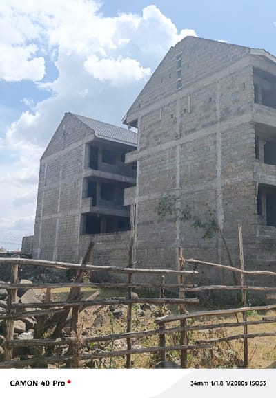 The image depicts an outdoor scene in Nakuru East ward, Kenya, featuring two multi-story buildings under construction. In the foreground, a rustic wooden fence, constructed from horizontal logs supported by vertical posts, spans the lower portion of the frame. The ground immediately behind the fence is dry, rocky, and covered with sparse, dry vegetation. The central subjects are two unfinished structures, primarily built with grey concrete blocks. The building on the left, set further back, displays a grey tiled pitched roof and multiple unglazed rectangular openings across its visible three-story height. The building on the right, positioned closer, features a partially tiled pitched roof and exposed concrete block walls with visible structural columns and numerous large, unglazed rectangular openings across its four discernible levels. A small, slender tree with sparse green leaves stands between the fence and the rightmost building. The background shows a blue sky with scattered white cumulus clouds. No individuals, specific actions, or direct interactions are visible.