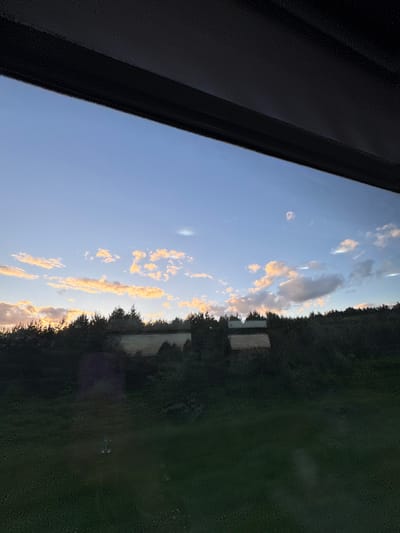 A sideways view from inside a building, possibly through a window with condensation on the glass. To the left is a black frame, likely part of the window structure. The view outside shows a blue sky with scattered, sunlit clouds. On the right side of the frame, there is greenery, likely trees or shrubs, and what appear to be white buildings or structures partially obscured by the foliage. The location appears to be a rural or suburban area in Мажэйкаўскі сельскі Савет, Belarus. The scene is bathed in the warm light of either sunrise or sunset.