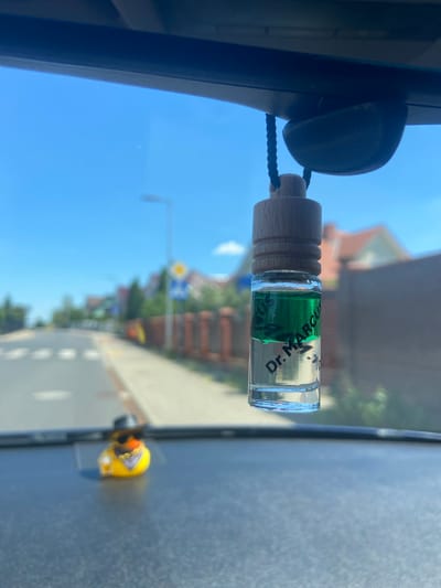 The image shows a car's interior, with a clear blue sky and houses visible through the windshield. A small glass bottle with green liquid and a wooden cap hangs from the rearview mirror. The bottle has text that reads "Dr. Marcus." In the foreground, on the dashboard, is a small yellow duck figurine wearing sunglasses and a bandana. The scene appears to be on a street in Bolesławiec, Poland, indicated by the road markings and houses.