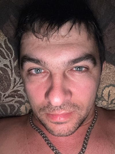 A close-up, head-on, selfie-style shot features a man with dark hair, light blue eyes, and a light beard. He is wearing a silver chain around his neck. The background consists of a patterned surface, possibly a pillow or blanket. The lighting is dim, creating a warm glow on his skin. There is no discernible location within the city of Shakhty, Russia, depicted in this image; it appears to be an indoor scene.