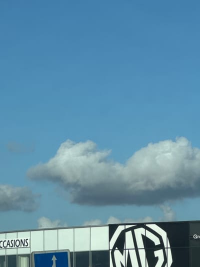 The image depicts an outdoor daytime scene in Souffelweyersheim, France, dominated by a clear blue sky with scattered cumulus clouds occupying the upper two-thirds. A prominent, elongated cloud mass stretches across the mid-ground. In the lower third, a modern commercial building is visible, featuring a facade composed of light-colored panels separated by vertical mullions. On the left side of the building, the partial word "CCASIONS" is discernible in white lettering on a dark background. The right side of the building prominently displays a large, black and white octagonal "MG" logo. To the immediate right of the MG logo, the partial word "Gro" is visible. In the foreground, partially obscuring the building, is a blue rectangular road sign with a white upward-pointing arrow. No people are visible, and no actions or interactions are occurring. The environment suggests a commercial or automotive dealership setting.