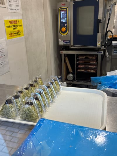 The image shows a commercial kitchen in Chiyoda City, Japan (latitude: 35.681, longitude: 139.768). A large, stainless steel oven with a digital display is mounted on a wall. The oven's door is open, revealing a rack of stacked trays containing food. To the right of the oven, there is a large white tray holding several plastic containers of food, each containing a green leafy vegetable. A blue plastic sheet is also visible on the counter. A yellow sign with Japanese text is attached to the wall near the bottom of the image. The sign is likely a warning sign. The containers of food are likely for sale or display. The kitchen appears to be clean and organized. There are no people visible in the image.