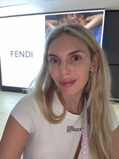 A woman with blonde hair and fair skin is taking a selfie in front of a Fendi advertisement at Fiumicino, Italy. She is wearing a white ribbed t-shirt. The advertisement behind her features the Fendi logo and a partial image of a person with red hair. The floor appears to be tiled.