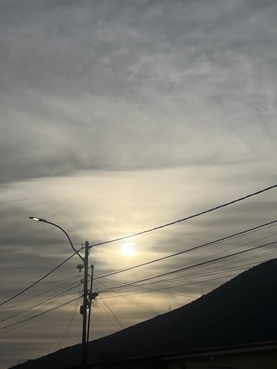 The image shows a view of a cloudy sky with the sun partially visible behind the clouds, casting a diffused light. In the foreground, a silhouette of a steep mountain slope dominates the lower right portion of the frame. A utility pole with numerous wires is centrally located, with a streetlamp extending from the left side of the pole. The wires are strung across the image, creating a dense network. In the very bottom of the frame, a sliver of a building's roof is visible. The overall scene suggests an urban or semi-urban environment with infrastructure and natural elements coexisting. The location is identified as San Antonio, Venezuela, but no specific landmarks are discernible to confirm this without external information. There are no people or discernible actions occurring in the image.
