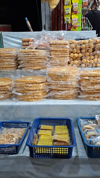 In a market stall in Keudegeudong, Indonesia, a variety of packaged snacks are displayed for sale. The stall features a densely packed array of items, with many hanging from above and others arranged in blue plastic baskets.
Prominently displayed are numerous clear plastic packages containing small, round, golden-brown snacks, possibly puffed rice or a similar confection. Adjacent to these are packages of elongated, flat snacks, also golden-brown and appearing to be made of seeds or grains. A few larger, irregularly shaped snacks are visible in clear bags on the left.
In the blue baskets, different types of snacks are organized. One basket holds stacks of rectangular, layered snacks, predominantly yellow with some brown ones interspersed. The lower basket contains a collection of light brown, irregularly shaped crispy snacks, possibly chips or crackers.
A person is partially visible in the upper left corner, wearing a dark shirt, suggesting they are the vendor or a customer. The background is a pale, textured wall, with shadows cast on it, indicating indoor lighting. The overall impression is of a bustling market stall focused on selling a wide range of snack foods.
Visible text includes brand names and product descriptions on some of the snack packages. For instance, yellow packages have the word "POPCORN" and "ATOM" clearly printed. Another package has Indonesian text, and a small yellow label with text is visible on one of the large snack packages. The overall scene conveys a sense of commerce and readily available food items.