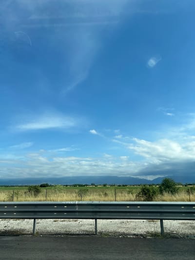 The image captures a roadside scene on a bright, partially cloudy day in Malo Konare, Bulgaria. The primary subject is a multi-tiered metal guardrail, dark grey with reflective horizontal strips, stretching across the middle ground. To the right of the guardrail, a dark asphalt road surface is visible, bordered by a lighter-colored gravel and dirt shoulder. On the guardrail itself, faint, light-colored markings, possibly graffiti, are visible, appearing to spell out "COOL" in the central area.
Beyond the guardrail, to the left, lies an expansive agricultural field with dry, yellowish-green grass and scattered trees and shrubs. In the far distance, under a hazy sky with more substantial cloud cover, the faint silhouettes of mountains or large hills are discernible.
The sky occupies the upper portion of the image, showcasing a vibrant blue with various white cloud formations. These range from thin, wispy cirrus clouds to larger, fluffier cumulus clouds, particularly towards the distant horizon. Several circular and semi-circular lens flare artifacts are present in the upper left quadrant of the sky. The overall impression is one of a calm, open rural landscape viewed from a moving vehicle. No people or specific activities are depicted beyond the implied travel. The time of day appears to be mid-day due to the bright, well-lit conditions.