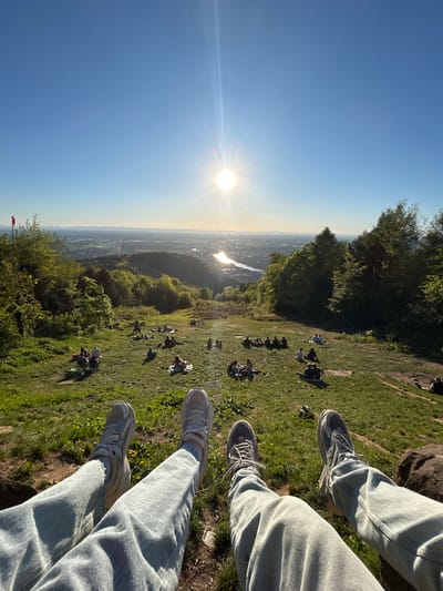 The image shows a viewpoint on the Königstuhl hill in Heidelberg, Germany. The photo is taken in a vertical perspective from a high vantage point, likely where someone is sitting with their legs dangling over the edge. Two pairs of legs wearing jeans and sneakers are visible in the foreground. Below, a grassy slope is dotted with people lounging and sitting in groups. The city of Heidelberg is visible in the background under a clear, sunny sky, with the sun glaring brightly. Trees line the edge of the hill and dot the landscape. A red and white windsock is faintly visible at the very bottom left corner.