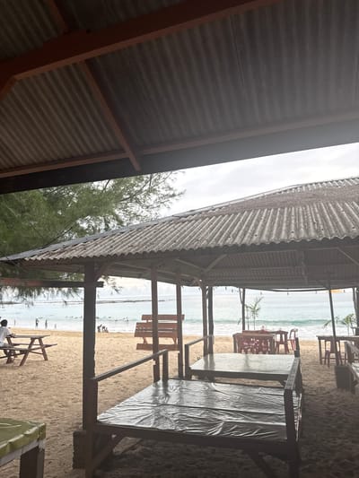 This image captures a beach scene in Meunasahbale, Indonesia, viewed from under a corrugated metal roof structure. The setting appears to be a beachside cafe or restaurant with tables and benches arranged on the sand. In the foreground, part of the roofing structure with its corrugated metal sheets and wooden beams is visible.
Beyond the shelter, a sandy beach stretches towards the ocean. The water is a light blue-green, and there are small waves. Several people are scattered on the beach and in the shallow water, suggesting a leisurely day. Some appear to be swimming or wading, while others are simply walking along the shore. In the middle ground, more tables and chairs are set up, some with red plastic seats. Palm trees are visible, adding to the tropical ambiance.
The sky is overcast, indicating a cloudy day, possibly early morning or late afternoon given the soft lighting. The overall mood is relaxed and tranquil. There is no visible text in the image. The perspective is from inside a shaded area, looking out towards the open beach and sea. The orientation of the image is sideways, with the horizon running vertically.