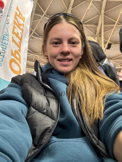 A woman with long, light brown hair, wearing a blue hooded sweatshirt and a black puffer vest, is looking at the camera and smiling. She has a black headband on. Behind her is the ceiling of a large indoor space with a white and metal support structure. In the lower left corner, there is a white banner with orange lettering that reads "ОБЪЕКТЫ". This image was taken in the city of Песчаные Ковали, Russia.