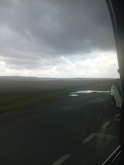 The image captures an expansive, open landscape within Malewa West ward, Kenya, viewed from the interior of a vehicle. The upper half of the frame is dominated by a dense, dark grey overcast sky, with a localized lighter patch of sky featuring white clouds visible towards the right horizon. A distinct, low-lying dark ridge or range of hills extends across the distant horizon, separating the sky from the terrain below. The midground and foreground consist of flat, open land. The left portion of this land exhibits dark green vegetation. The right portion features dark, wet ground with multiple irregular puddles of standing water reflecting the overcast sky. Numerous small, light-colored specks are scattered across this wet ground, consistent with the appearance of birds or debris. The extreme right edge of the frame shows a dark vertical vehicle structure, likely a window pillar. A vehicle side mirror is visible in the mid-right, reflecting elements of the sky and landscape. Indistinct reflections are present on the vehicle's window in the lower right quadrant. No human subjects or discernible actions are present. The scene indicates an environment characterized by recent or ongoing wet conditions.